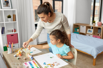 family, motherhood and leisure concept - mother spending time with her little daughter drawing or painting wooden chipboard items with colors at home