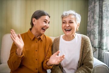 Elderly sisters watching comedy together indoors, laughing. Portrait of two beautiful Caucasian women relaxing at home, sitting on sofa, opening mouth wide opened, enjoying weekend, having fun - 431443075