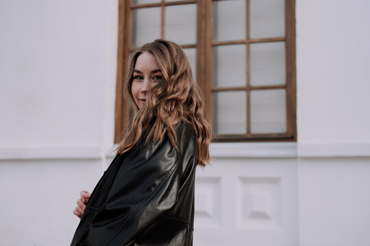 Smiling Young Woman In Black Leather Jacket Standing In Front Of White Wall Looking At Camera Over Shoulder. Urban Style And Street Fashion. Girl With Curly Hairstyle In Casual Outfit