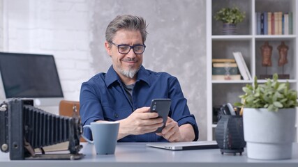 Bearded man working online with laptop computer and smart phone at home sitting at desk. Businessman in home office. Portrait of mature age, middle age, mid adult man in 50s.