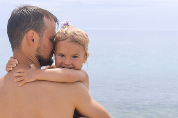 Portrait of child and dad. Cute little caucasian child hugs her young dad, father holds his kid and kisses, sea at background, happy childhood, good relationship between father and daughter concept.