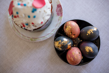homemade colored Easter eggs with gold ornaments in a black plate on a gray cloth and Easter cake with white glaze in the background. Spring, religious fast, Easter holiday, traditions