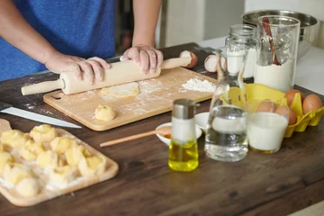 Foto op Canvas Female hands roll out a piece of dough with a rolling pin for making ravioli or dumplings on a wooden table sprinkled with flour. A step-by-step recipe for cooking ravioli or dumplings © Cuong