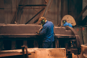 Worker in workshop processes large cast iron part after casting in metallurgical foundry plant.
