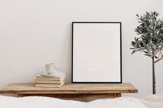 Vertical black picture frame mockup on vintage bench, table. Cup of coffee on pile of books. Potted olive tree. White wall background. Scandinavian interior, neutral color palette. Selective focus.