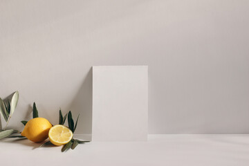 Summer stationery still life scene. Cut lemon fruit and olive tree branch on white table background in sunlight. Blank vertical greeting card mockup leaning against beige wall. Branding concept.