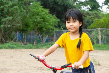 Fototapeta na wymiar happy little cute Asian girl dress casually standing holding a bicycle handle looking at a camera in a countryside farm of northern Thailand natural background. Healthy child and holiday concept