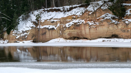 Fototapeta na wymiar landscape with frozen river Gauja near Gudu cliffs, view of sandstone cliff from the opposite bank of the river, beautiful red rock wall, bare trees on the river bank, winter, Ligatne, Latvia