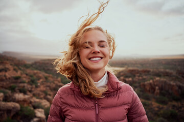 Smiling young confident woman with closed eyes feeling fresh wind against face standing on mountain...