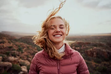 Fotobehang Smiling young confident woman with closed eyes feeling fresh wind against face standing on mountain hill © StratfordProductions