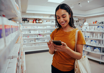 Young woman smiling while using smartphone standing against shelf in pharmacy