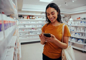 Poster Young woman smiling while using smartphone standing against shelf in pharmacy © StratfordProductions