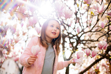 Preschool girl in a grey dress wearing rosa jacket staying under a flowering pink magnolia tree