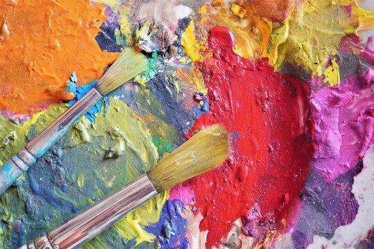 Close up of two paintbrushes with bristles, colorful mixed paints and grunge style painted palette.  Bright multi-colored background image and creative concept.
