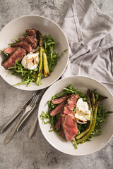 Gerösteter grüner Spargel mit medium rare Rinder Steak Streifen, Rucola, Büffel Mozzarella, Olivenöl und Balsamico in Schale mit Leinen Serviette, Besteck und rustikal Stein Hintergrund
