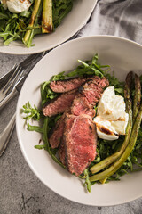 Gerösteter grüner Spargel mit medium rare Rinder Steak Streifen, Rucola, Büffel Mozzarella, Olivenöl und Balsamico in Schale mit Leinen Serviette, Besteck und rustikal Stein Hintergrund
