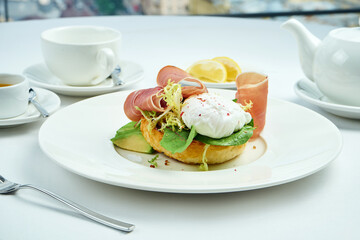 Delicious breakfast - toast with avocado, jamon crudo and poached egg, served in a white plate on a white tablecloth. Close up, selective focus