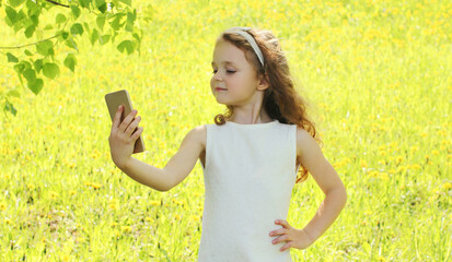 Naklejka na ściany i meble Portrait of little girl child taking a selfie by smartphone in summer park