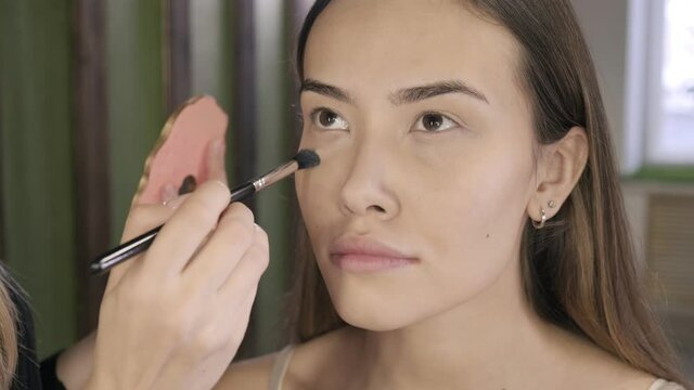 closeup on hand of make up artist doing makeup for young brunette asian woman with brush in beauty salon. visagiste working, preparing female client for event or photoshoot, applying concealer on face