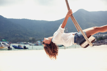 Young beautiful woman in a white shirt swinging on a swing over the sea, summer travel and vacations - obrazy, fototapety, plakaty