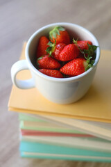 Mug filled with fresh strawberries and stack of book on a table. Selective focus.
