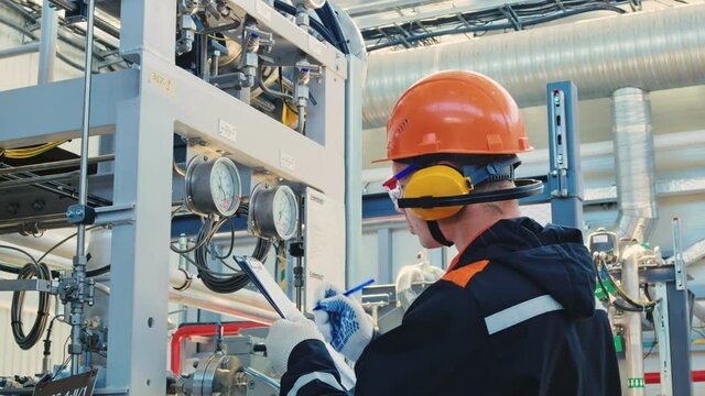 An engineer technician in a hard hat, goggles and headphones, writes the data from a pressure gauge and a tremometer with a pen on a sheet of paper. Work in industrial. Manufacturing industry.