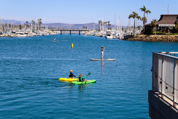 a woman standing on a paddle board with two kids in green and yellow kayaks on deep blue ocean water with boats and yachts docked in the harbor at Baby Beach in Dana Point California - obrazy, fototapety, plakaty