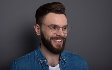Confident smart modern bearded man in casual denim clothes and eyewear is posing over gray background in the studio