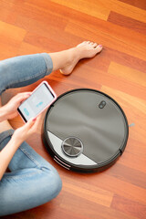 Young girl feet at home using a smart phone to control a round smart automatic vacuum, machine...