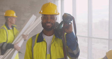 Portrait of smiling afro-american builder in uniform holding drill on his shoulder