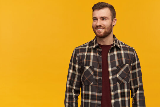 Smiling attractive young man in plaid shirt with beard standing and looking away to the side over yellow background
