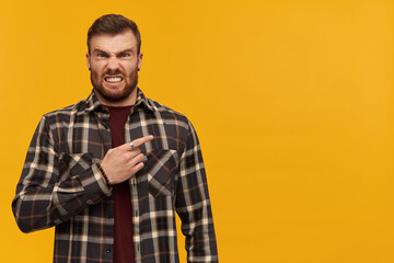 Angry irritated young man in plaid shirt with beard standing and pointing by finger away to the side over yellow background