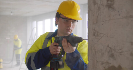 Builder in helmet and glasses with electric drill making hole in wall