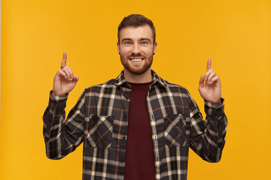 Cheerful charming young man in checkered shirt with beard standing and pointing up to the sky with two fingers over yellow background