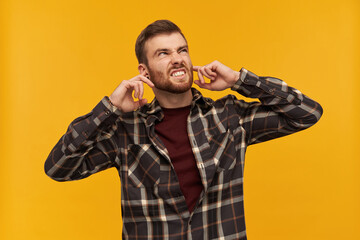 Annoyed angry young man in checkered shirt with beard covered ears by fingers and feels irritated...