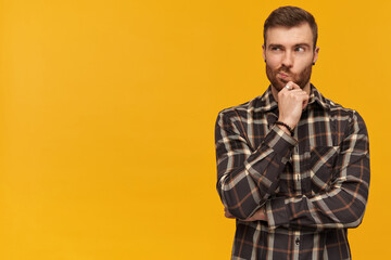 Pensive handsome young bearded man in checkered shirt touching his chin and thinking over yellow...
