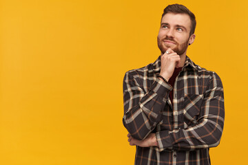 Smiling pensive young man in checkered shirt with beard dreaming and looking away to the side over yellow background Looks inspired