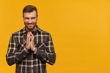Mischievous arrogant young man in plaid shirt with beard rubbing hands and scheming an evil plan over yellow background Looking away to the side