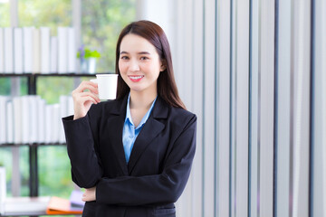 Asian business lady who wears blue shirt and black suit holds coffee cup in her hand and smile happily at work office as background.