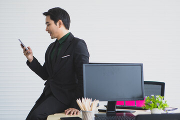 A young man in black suit, dark eyebrows, charming eyes. Looking smiling at the camera talking on phone Sit and rest on desk. Looks professional warm and easy going at the same time.