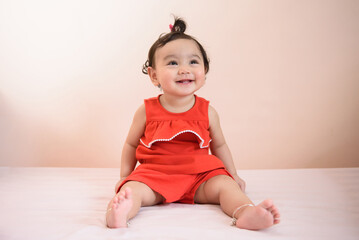 Portrait of a smiling cute little Asian baby girl wear red dress siting on bed and looking out isolated on background, baby expression concept