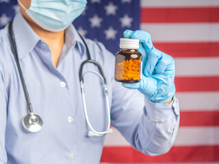 Healthcare and medical concept. Doctor wearing a face mask, hand wearing blue gloves, and holding a pills bottle against the background of the United States flag