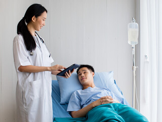 Obraz premium In hospital private room, One female doctor with stethoscope and laptop in her hands talking with patient man as he laying on bed with IV tube. Look each other with smile. Seems guy recovery soon.