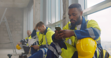 Diverse builders at construction site resting during break using smartphone