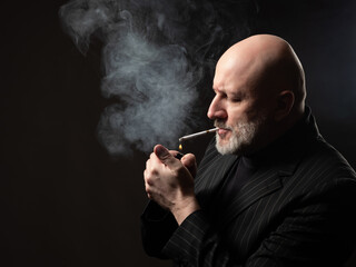 The man lights a cigarette. Man with a lighter and a cigarette. A smoker in a dark suit against the...