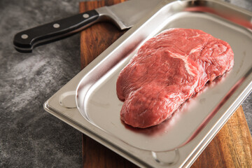 Rinder Steak roh auf Edelstahl Schale mit Küchenmesser, Holzbrett und Stein Hintergrund dunkel