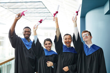 Waist up view at diverse group of cheerful young people wearing graduation gowns and smiling at...