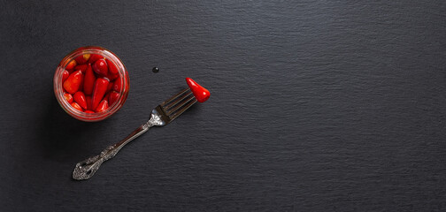 Open glass jar of pickled red hot peppers and one more on the fork against black slate background. Wide angle image for food design mockup with fermented vegetables. Copy space.