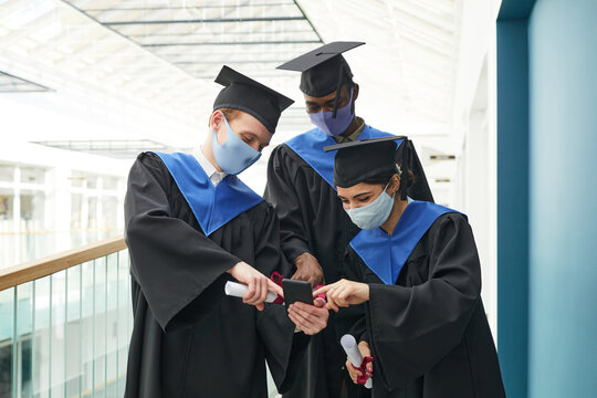 Diverse group of young people wearing graduation gowns and masks taking selfie indoors in college interior, copy space