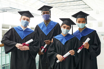 Diverse group of young people wearing graduation gowns and masks while posing indoors in college interior